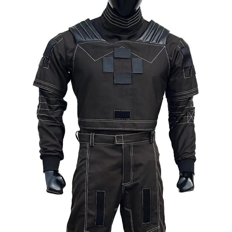 May include: Black tactical jumpsuit with white stitching details. The outfit features a high collar, shoulder accents, and multiple square patches on the chest. The long-sleeved top and pants are connected by a belt.