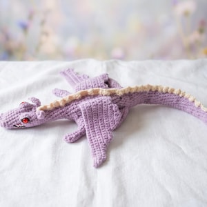 May include: A handmade crochet dragon in lavender with cream accents. The dragon has red eyes and detailed wings. The toy is lying on a white surface, with a soft, blurred background.