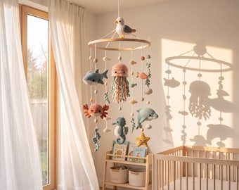 May include: A hanging mobile featuring a circular wooden frame with a crocheted seagull on top. Suspended from the frame are various crocheted sea creatures including a shark, jellyfish, crab, seahorse, dolphin, and starfish, interspersed with small wooden beads.