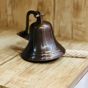 May include: A polished dark bronze wall-mounted bell featuring a curved metal bracket for attachment and a thick, braided off-white rope pull attached to the clapper, designed for clear sound projection in a nautical or functional setting.