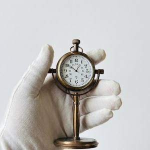 Può includere: Un orologio da tavolo color ottone con quadrante bianco rotondo, numeri neri da 1 a 12 e un anello interno con indicatori delle ore da 13 a 24. L'orologio è montato su un supporto verticale con base circolare e una manopola di carica in alto.