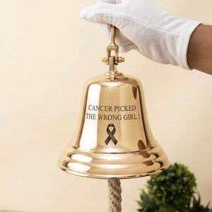 Könnte beinhalten: Eine polierte goldfarbene Messingglocke mit einem dicken geflochtenen Seilgriff. Die Glocke ist mit dem Text CANCER PICKED THE WRONG GIRL! graviert und enthält ein schwarzes Awareness-Schleifen-Symbol, das unter dem Text auf der Metalloberfläche zentriert ist.