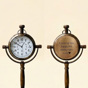 May include: A dual-sided antique-style brass desk clock on a stand. One side features a white clock face with black numerals 1-12 and 13-24, while the other side displays the engraved text: 6 years of love happy 6th anniversary to us.