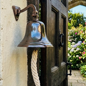 Puede incluir: Una campana de pared de bronce pulido con un soporte de montaje curvo y un tirador de cuerda trenzada de color blanco roto. La campana tiene una superficie lisa y reflectante y está fijada a una pared clara junto a una puerta de madera oscura.