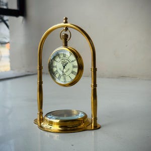 May include: A polished brass desk clock suspended within an arched frame, featuring a round face with Roman numerals, black hands, and the text Victoria London 1876. A circular compass is integrated into the base of the brass stand.
