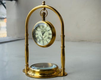 Op de afbeelding: Een gepolijste messing bureauklok hangend in een boogvormig frame, met een ronde wijzerplaat met Romeinse cijfers, zwarte wijzers en de tekst Victoria London 1876. Een rond kompas is geïntegreerd in de voet van de messing standaard.
