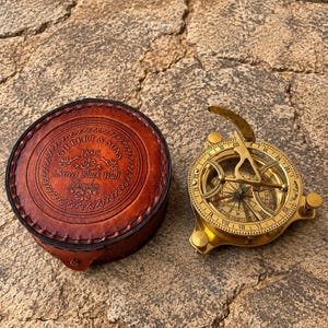 May include: A round brown leather case with embossed text reading Gilbert & Sons 1895 8 Street Black Wall London, paired with a circular brass sundial featuring a folding gnomon, Roman numerals, and three adjustable leveling screws on the base.