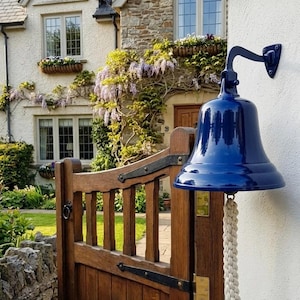Puede incluir: Una campana de metal azul oscuro brillante montada en una pared blanca con una cuerda gruesa trenzada de color blanco roto que cuelga del centro. La campana cuenta con un soporte de metal curvado fijado a la pared, creando un elemento funcional.
