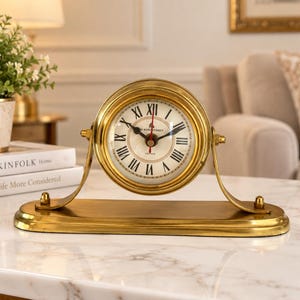 May include: A gold-toned metal desk clock with a circular face, black Roman numerals, and black hands. The clock is mounted on a curved, arched stand attached to a rectangular base. The clock face features the text 89 KENSINGTON LONDON.