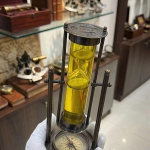 May include: An antique brass hourglass with a compass base. The hourglass contains yellow sand and is held by a gloved hand. The design features a nautical theme, with a compass rose visible at the base. The background includes other nautical items.