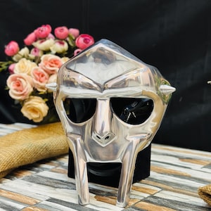May include: A polished silver mask with a distinctive, angular design. The mask features a prominent nose, eye openings, and sharp, conical spikes on the sides. The mask is displayed on a black surface, with a bouquet of pink and cream roses in the background.