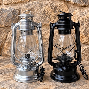 May include: Two hurricane lanterns, one in a polished silver finish and one in a matte black finish. Each lantern features a clear glass chimney, a protective wire cage, a top handle, and a black electrical cord extending from the base.