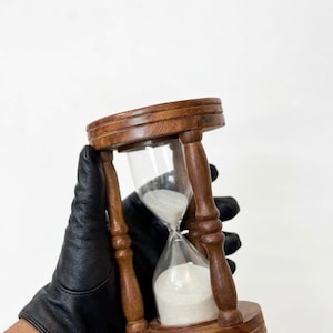 May include: A wooden hourglass with a clear glass center filled with white sand. The hourglass is held by a person wearing a black glove. The wooden frame is a dark brown color.