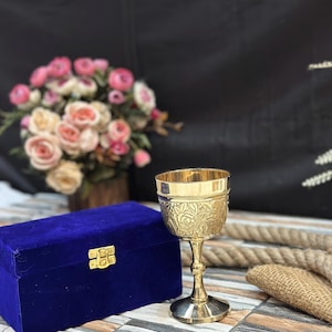 May include: A gold-colored chalice with floral engravings, set next to a blue velvet box with a gold clasp. A bouquet of pink and cream roses is in the background.