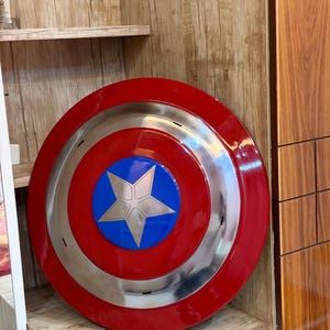 May include: A circular shield featuring a glossy red outer ring, a metallic silver middle ring, and a bright blue center circle containing a large, five-pointed silver star. The shield has a smooth, reflective finish and a convex shape.
