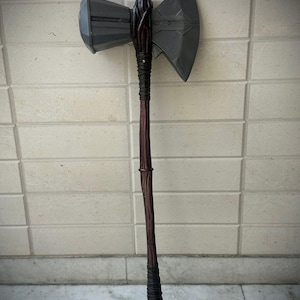 May include: A large battle axe featuring a dark grey metallic head with a hammer-like side and a sharp blade side. The handle is long, brown, and textured to resemble wood, with dark wrapped sections at the top and bottom of the shaft.