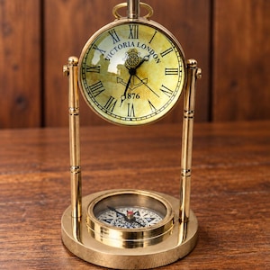 May include: A brass desk clock and compass set featuring a round clock face with Roman numerals, the text Victoria London, and an anchor graphic. The clock is suspended between two vertical brass posts above a circular base with a compass.