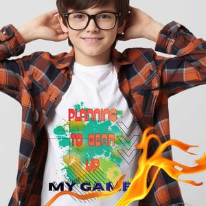 May include: White t-shirt featuring the text "PLANNING TO GEAR UP MY GAME" in a colourful, graffiti-style design. The shirt is worn with a plaid button-down shirt and glasses.