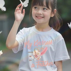 May include: A young person wearing a white t-shirt with the text "My Kindness carries me like a Feather" in peach-colored script. The shirt features a heart and feather design. The person is smiling and surrounded by white butterflies.