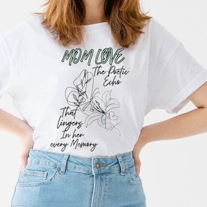 May include: White t-shirt with the text "MOM LOVE The Poetic Echo That lingers In her every Memory" in green and black. The shirt features a floral design. The model is wearing light blue jeans.