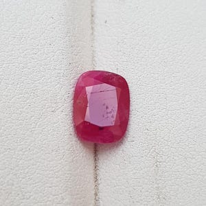 May include: A cushion-cut ruby gemstone with a deep red hue. The gemstone has a faceted surface that reflects light, and is set against a white background. The ruby is a vibrant and eye-catching piece.