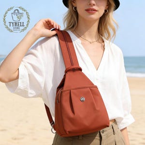 Puede incluir: Un bolso bandolera de color óxido con una sola correa para el hombro. El bolso tiene un compartimento principal con cremallera, un pequeño bolsillo frontal y un emblema circular plateado. El bolso se lleva sobre el hombro.