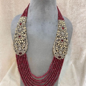 Puede incluir: Collar de múltiples hilos en un tono rojo intenso, con colgantes dorados ornamentados adornados con piedras preciosas claras y rojas. El collar se exhibe en un maniquí gris, mostrando su intrincado diseño.