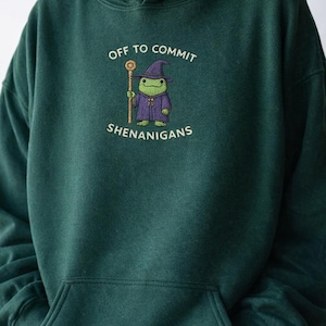 May include: Dark green hoodie with a front pocket and a drawstring hood. The front features the embroidered text "OFF TO COMMIT SHENANIGANS" above a cartoon frog wearing a wizard hat and robe, holding a staff.