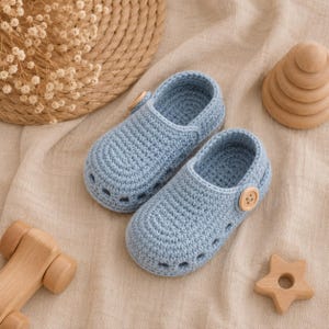 May include: A pair of light blue crochet baby shoes featuring a rounded toe, side cutouts, and a single wooden button on the outer side of each shoe. The shoes are crafted with a textured yarn stitch and have a slip-on design with a heel strap.
