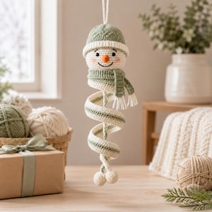 May include: A crocheted snowman ornament featuring a sage green hat and matching scarf, with a spiral-shaped body extending downward. The snowman has a white face, a small orange carrot nose, and two small white pom-poms at the base of the spiral.