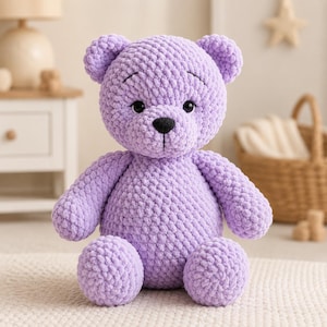 Peut inclure: Un ours en peluche fabriqué en fil violet clair avec un motif de mailles bouclées texturées. L'ours présente deux oreilles rondes, de petits yeux en plastique noir et un nez noir brodé. Il est assis en position verticale, les bras le long du corps.