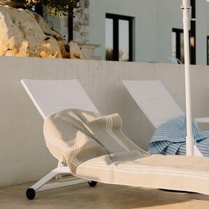 Può includere: Due sedie a sdraio bianche con asciugamani a righe beige e blu. Le sedie sono su un patio di cemento, con un edificio bianco e un albero sullo sfondo. È visibile anche un ombrellone bianco.