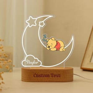 May include: A night light featuring a sleeping Winnie the Pooh on a crescent moon with stars. The lamp has a wooden base with the text "Custom Text" and a cloud with the name "Naylar".