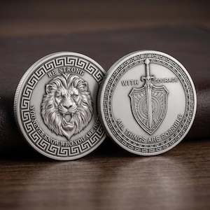 May include: Two circular silver-toned metal coins featuring embossed designs. One coin displays a roaring lion head with the text BE STRONG YOU NEVER KNOW WHO YOU ARE INSPIRED. The other coin shows a sword and shield with WITH COURAGE ALL THINGS ARE POSSIBLE.