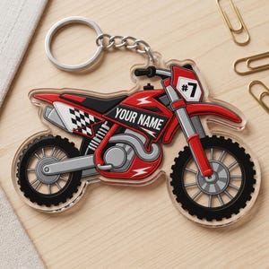 Pode incluir: Um porta-chaves de acrílico transparente em forma de mota de cross vermelha com pneus pretos, uma placa branca com o texto #7 e um painel lateral com o texto YOUR NAME. O design da mota inclui um padrão de xadrez preto e branco.