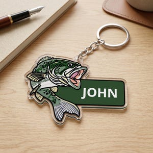 May include: A clear acrylic keyring featuring a colourful illustration of a largemouth bass fish with its mouth open, attached to a silver metal split ring. Next to the fish is a dark green rectangular tag with the white text JOHN printed on it.