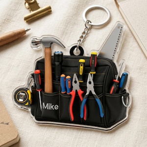 Puede incluir: Un llavero de acrílico transparente con forma de bolsa de herramientas negra con costuras amarillas. La bolsa contiene un martillo, linterna, destornilladores, alicates, una sierra y una cinta métrica. El nombre Mike está impreso en blanco.