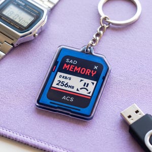 May include: A rectangular acrylic keychain shaped like a memory card with a blue border and a white center screen. The screen displays the text SAD MEMORY, 0 KB/S, 256MB, and ACS, along with a sad face icon. A silver metal chain and ring are attached.
