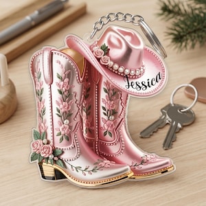 Op de afbeelding: Een roze acryl sleutelhanger met een paar cowboylaarzen en een cowboyhoed. De laarzen en hoed zijn versierd met roze bloemmotieven, kleine witte parels en groene bladeren. De naam Jessica staat in zwarte letters op de hoed gedrukt.