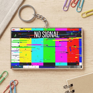 May include: A rectangular acrylic keychain featuring a television test pattern with vertical bars in yellow, cyan, green, magenta, and red. The design includes digital glitch effects and the white text NO SIGNAL printed across the upper section.