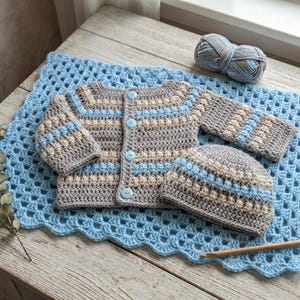 Pode incluir: Um conjunto de bebê de crochê feito à mão em cinza, azul e bege, incluindo um cardigã com botões, um gorro e um cobertor. O conjunto é exibido em uma superfície de madeira com uma bola de lã e uma agulha de crochê. O cobertor tem uma borda ondulada.