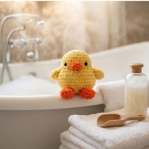 May include: A small, round, yellow crocheted duck toy with a bright orange beak and orange feet. The duck has two small black eyes and is positioned on the edge of a white bathtub. The texture of the yarn is clearly visible in the stitching.