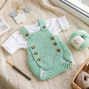 May include: A mint green crochet baby romper with wooden buttons, paired with a white long-sleeved shirt. A ball of mint green yarn, a crochet hook, and a small wooden mobile are also present, suggesting a handmade baby clothing theme.