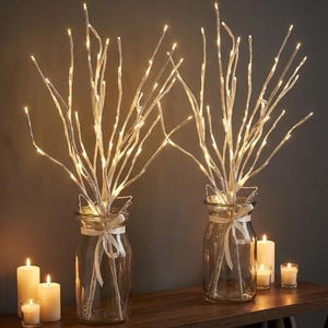 May include: Two glass jars filled with white, illuminated branches. Each jar is adorned with a white ribbon. Several lit candles of different sizes are on a wooden surface, creating a warm atmosphere.