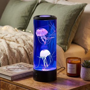 May include: A cylindrical jellyfish lamp with a black base and top. The lamp's blue interior houses two jellyfish figures with long, flowing tentacles. The jellyfish are pink and white. The lamp is on a wooden nightstand next to a candle and a succulent.