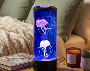 May include: A cylindrical jellyfish lamp with a black base and top. The lamp's blue interior houses two jellyfish figures with long, flowing tentacles. The jellyfish are pink and white. The lamp is on a wooden nightstand next to a candle and a succulent.