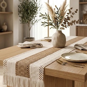 May include: A wooden dining table set with a cream-colored table runner featuring a woven jute center and fringe trim. A beige vase holds dried plants. The table is set with plates, napkins, and gold-colored cutlery.