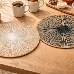 Könnte beinhalten: Zwei runde, gewebte Tischsets mit radialem Design. Eines ist beige und weiß, das andere schwarz und beige. Beide haben einen hellbraunen Rand. Sie liegen auf einem Holztisch, zusammen mit Kaffeetassen und einer Schüssel Erdbeeren.