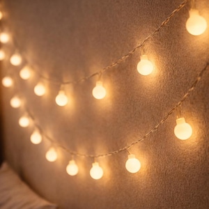 Könnte beinhalten: Warme, weiße Lichterkette mit runden Glühbirnen an einer strukturierten Wand. Die Lichter sind gleichmäßig verteilt und strahlen ein weiches, einladendes Leuchten aus, das eine gemütliche Atmosphäre schafft. Der Hintergrund ist ein neutraler, warmer Ton.