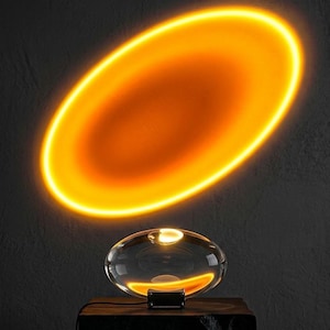 May include: A modern sunset lamp with a clear, oval-shaped glass body and a black base. The lamp projects a warm orange, oval-shaped light onto the wall, creating a sunset effect. The lamp sits on a dark wooden surface.
